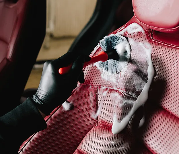 Main gantée nettoyant un siège de voiture en cuir rouge perforé avec une brosse et du savon à Maisons-Laffitte dans les Yvelines 78